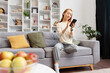 Young Woman Enjoying Time On Sofa At Home, Smiling at Smartphone in Cozy Living Room With Bookshelves, Comfort, Leisure, And Modern Lifestyle