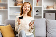 Young Asian Woman With Credit Card And Smartphone Sitting On Sofa, Smiling At Camera, Reflecting Online Shopping And Modern Lifestyle In Cozy Living Room.