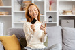 Happy Young Woman Using Smartphone And Holding Credit Card On Couch, Online Shopping, Mobile Banking In Modern Living Room.