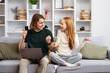 Young Couple Enjoying A Lively Conversation On Couch, Man Holding Credit Card And Laptop, Woman Gesturing With Hands, Cozy Home Background. Online Shopping, Casual Lifestyle, Modern Couple At Home.