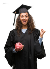 Poster - Young hispanic woman wearing graduation uniform holding piggy bank screaming proud and celebrating victory and success very excited, cheering emotion