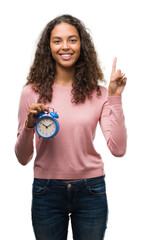 Wall Mural - Young hispanic woman holding alarm clock surprised with an idea or question pointing finger with happy face, number one