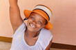 beautiful young african woman with a raised arm, wearing an orange hat sitting on the veranda in front of the house, township village in south africa