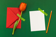 Red tulip taped on a green background, blank white card, red envelope and orange pencil.Greeting card, invitation.