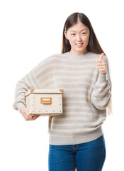 Sticker - Young Chinese woman over isolated background holding a box happy with big smile doing ok sign, thumb up with fingers, excellent sign