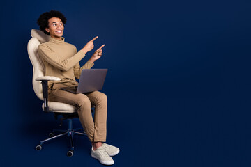 Full size photo of young man sit armchair netbook look point empty space wear trendy beige outfit isolated on dark blue color background