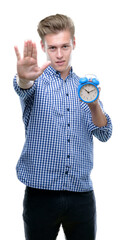 Canvas Print - Young handsome blond man holding alarm clock with open hand doing stop sign with serious and confident expression, defense gesture