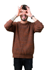 Wall Mural - Young handsome man wearing winter sweater over isolated background doing ok gesture like binoculars sticking tongue out, eyes looking through fingers. Crazy expression.