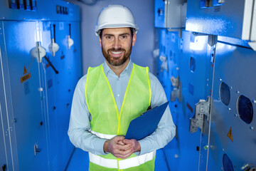 Positive factory worker in white hard hat and green safety vest hold clipboard. Electrician man looking at camera with happy face at electric technical blue room.