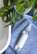 Spray cosmetic bottle on blue towels near basin and green monstera in bathroom, mockup