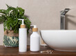 White one pump bottles near basin and green plant on wooden countertop in bath, mockup