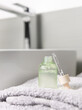 Opened cosmetic bottle with green serum on folded bath towel against basin and faucet close up