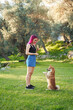 A woman with vibrant pink hair is training a Shiba Inu dog in a lush park, rewarding the attentive dog as it sits up on its hind legs