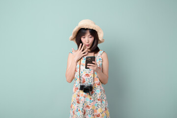 Happy Asian woman wearing casual dress and hat with camera while getting amazing news on mobile phone on vacation or travel theme isolated on pastel green background. Travel and vacation concept.