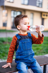 Sticker - A young boy is sitting on a bench drinking water. He is wearing a blue overalls and an orange shirt