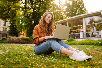 Wall Mural - Portrait of attractive young woman sitting on green grass in park while using laptop and wireless earphone. Freelancer, education, online, technology concept.