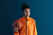 Portrait of young female basketball player against blue backdrop. Woman in orange sportswear holding a basketball looking at camera.