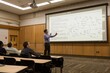 A man standing and giving a lecture in front of a large projection screen in a room, A professor giving a lecture in front of a large whiteboard filled with equations