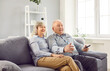 Portrait of smiling retired happy senior elderly couple fans watching football on tv holding TV remote in hands sitting on sofa in living room at home supporting sport team together.