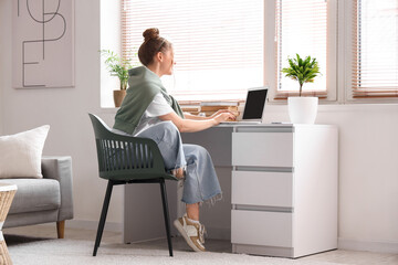Canvas Print - Young woman sitting at workplace and working with laptop in living room