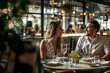 © Mau Martinez Cosio - A man and woman are sitting at a table in a restaurant, smiling at each other. The table is set with wine glasses, a fork, a knife, and a bowl. There are also flowers in vases and potted plants around