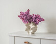 A bouquet of lilacs in a creative ceramic vase on a white chest of drawers