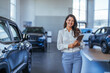 Elegant salesperson working at car dealership. Cheerful young woman with tablet PC smiling and looking at camera while working in modern car dealership. Saleswoman in car showroom selling cars