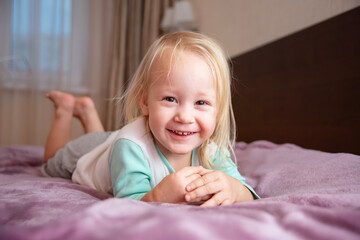 Portrait of a mischievous little girl. A little girl with shaggy white hair lies on the bed and smiles joyfully.