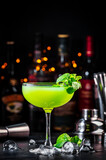 Fototapeta Na sufit - Summer green cocktail drink with gin, liqueur, lime, juice, mint and ice. Black bar counter background, steel bar tools and bottles