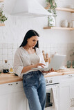 Fototapeta Do pokoju - Young woman freelancer or student working on a laptop in her kitchen, taking breaks between household chores or cooking. Perfect for content about balancing education, work, and home life