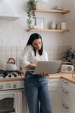Fototapeta Do pokoju - Confident young woman freelancer successfully balancing remote work and home chores, talk on the phone call and work on laptop in a well-organized, stylish kitchen. Concept of lifestyle management