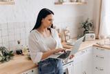 Fototapeta Do pokoju - Portrait of young woman freelancer or student working on a laptop in her kitchen, taking breaks between household chores or cooking. Perfect for content about balancing education, work, and home life