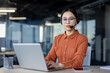 © Liubomir - Focused young Asian woman in a modern office environment. She wears a headset and glasses, looking at her laptop screen, exhibiting professionalism and concentration.