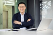 © Liubomir - A cheerful Asian male executive stands confidently at his desk in a well-lit, contemporary office space, radiating professionalism and positivity.