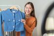 © Pormezz - social media market concept. Young Asian woman seller showing second-hand clothes during live streaming in the mobile application.