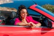 © svetograph - A woman in a pink jacket is sitting in a red convertible. She is wearing sunglasses and has her hand on her hip. Scene is relaxed and carefree.