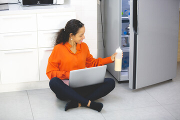 A Cheerful positive attractive African American is using laptop to learn online recipe, ingredient and cooking beside a refrigerator, Healthy lifestyle at home, Concept of home cooking preparation.