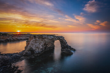 Wall Mural - Rocky coastline with a natural arch at punta Asparano, near Siracusa. Sunrise time, long exposure picture. June 2023