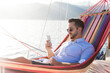 Man with mobile phone relaxing on yacht in hammock. Sea travel on sailboat. Traveler using smartphone and mobile app for banking, shopping online in summer vacation. Digital wellbeing, lifestyle