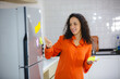 African female putting sticky note on refrigerator to remind her important job, work, plan, reminder, task and goal. Attractive smart Latin women putting or reading her note house activity checklist.