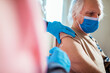 © Marko Geber - Senior man in face mask getting vaccinated at home