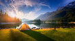 A tranquil morning at a mountain lake campsite. A tent sits near the calm waters, surrounded by lush greenery and towering peaks under the golden sunrise. Generative Ai.