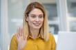 © Liubomir - Attractive blonde woman in a yellow blouse smiling and waving at the camera during a video call, sitting in a bright office space.