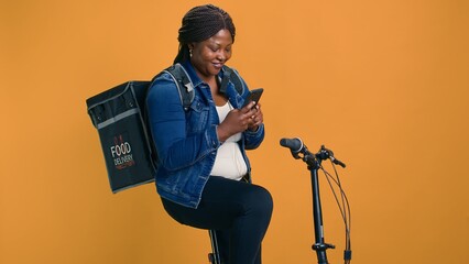 Healthy african american female with bag on her back using mobile communication for on-the-go urban delivery service. Active black woman happily receiving online orders on cell-phone from customers.