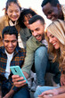 © CarlosBarquero - Vertical photo. Group of young people watching something at phone sitting on stairs. Smiling multicultural friends looking social media outside the university campus. Students in their free time.