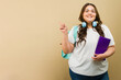 Smiling plus-size female student pointing towards copy space, ready for school with notebook