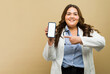 Cute Hispanic fat female doctor pointing at a blank smartphone screen in a beige studio setting showing a mobile app