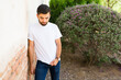 Latino man poses against a wall, looking down, showcasing a plain white t-shirt ideal for mockup designs