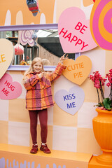 A young girl in an orange jacket standing in front of a colorful love-themed decoration with playful gestures.