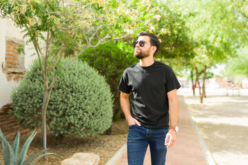 Wall Mural - Confident young hispanic man poses outdoors in a casual urban setting, wearing a trendy black t-shirt and stylish sunglasses